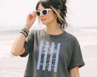 Camiseta cristiana "Paz, cálmate" con versículo bíblico de Marcos 4:39. Ropa holgada, top corto, ideal para vacaciones en la playa o como pareo. Diseño gráfico costero.