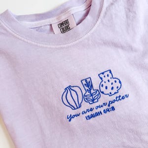 May include: A light purple t-shirt featuring blue embroidery of three distinct pottery vessels. Below the vessels, the text reads "You are our potter" and "ISAIAH 64:8". The inner neck label displays the brand name "COMFORT COLORS".
