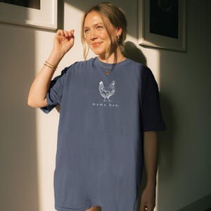 May include: A navy blue t-shirt with a white graphic of a hen and the text "mama hen." The shirt is made of a soft material and has short sleeves. The t-shirt is a comfortable, casual style.