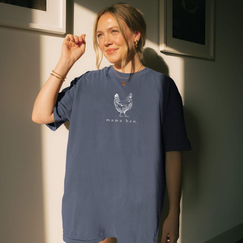 May include: A navy blue t-shirt with a white graphic of a hen and the text "mama hen." The shirt is made of a soft material and has short sleeves. The t-shirt is a comfortable, casual style.