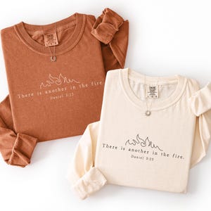 May include: Two long-sleeved shirts, one in a warm brown and the other in cream, are displayed. Each shirt features the text "There is another in the fire. Daniel 3:25" with a flame graphic. A silver necklace with a sun pendant is on each shirt.