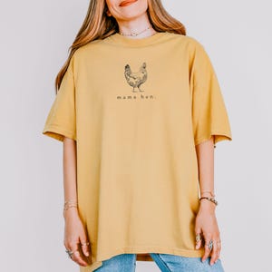 May include: Mustard yellow t-shirt with a black illustration of a hen and the words "mama hen." The shirt has short sleeves and a relaxed fit. The model is wearing blue jeans.