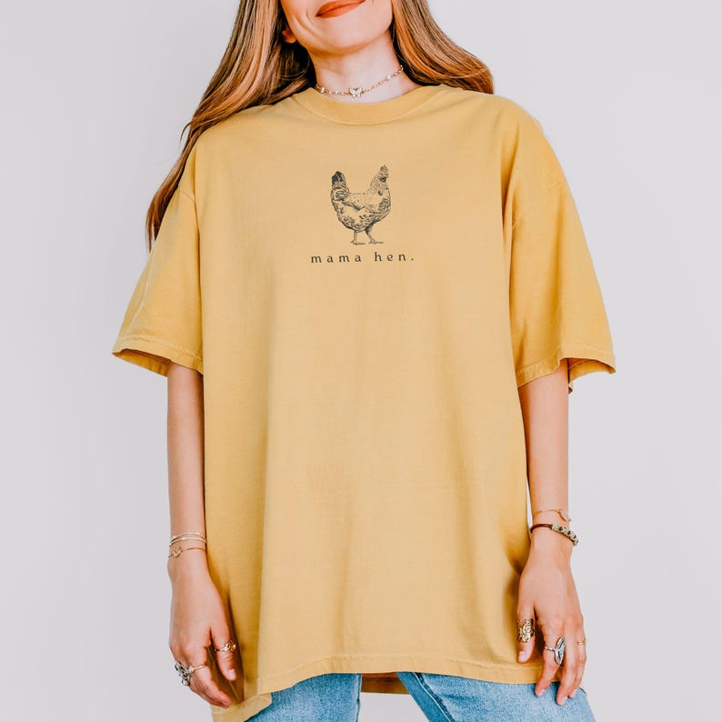 May include: Mustard yellow t-shirt with a black illustration of a hen and the words "mama hen." The shirt has short sleeves and a relaxed fit. The model is wearing blue jeans.