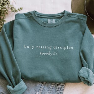 May include: Teal blue sweatshirt with the text "busy raising disciples proverbs 22:6" in white.