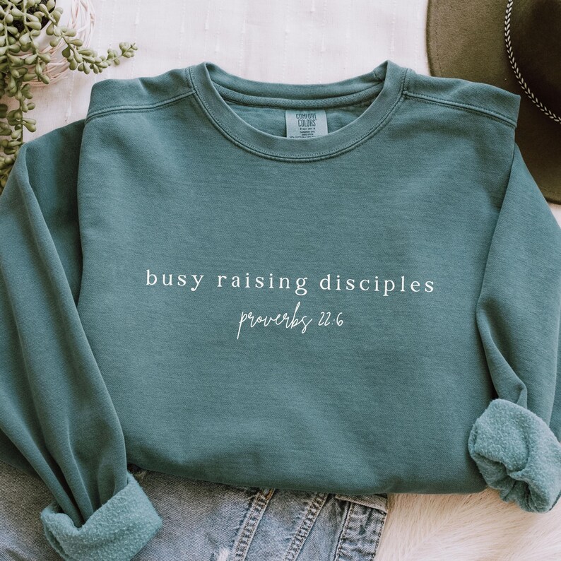 May include: Teal blue sweatshirt with the text "busy raising disciples proverbs 22:6" in white.