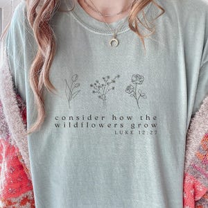 May include: Sage green t-shirt with a floral design and the text "consider how the wildflowers grow" and "LUKE 12:27". The shirt features three line-drawn flower illustrations.