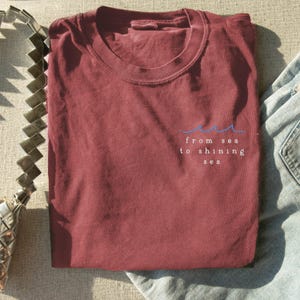 Könnte beinhalten: Ein maroonfarbenes T-Shirt mit einer weißen Grafik, auf der "from sea to shining sea" steht, mit einer blauen Wellenlinie über dem Text.