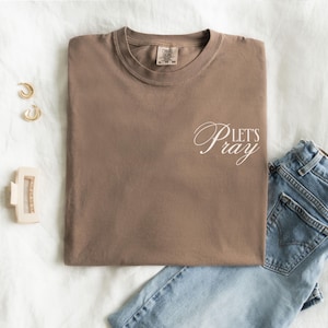 May include: A brown t-shirt with the words "Let's Pray" in white script. The shirt is folded neatly on a white surface, next to a pair of blue jeans, gold hoop earrings, and a hair clip.