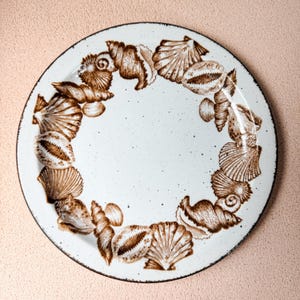 May include: A round ceramic plate with a white speckled finish and a dark rim. The plate features a circular border pattern of various brown seashell illustrations, including scallops, conch shells, and spiral shells arranged around the center.