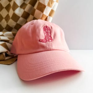 Pu&ograve; includere: Un berretto da baseball rosa con un designo di stivale da cowboy rosa ricamato.