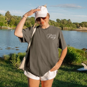 May include: A gray t-shirt with the text "Let's Pray" in a cursive font. The shirt is worn with white shorts and a white baseball cap. The background includes a body of water and green foliage.
