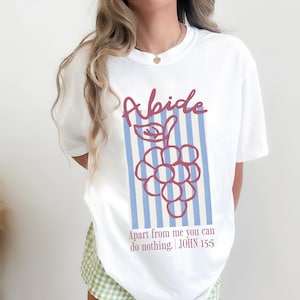 May include: White t-shirt with a graphic of a bunch of grapes and the word "Abide" in a script font. The design includes blue vertical stripes and the text "Apart from me you can do nothing. JOHN 15:5."