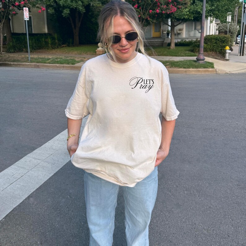 May include: Cream-colored t-shirt with the text "Let's Pray" in a script font. The shirt is paired with light blue jeans. The person is wearing sunglasses and a gold bracelet. The image is taken outdoors on a sunny day.