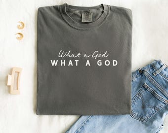 May include: A charcoal grey t-shirt with the words "What a God" in white script. Beneath, the same phrase is repeated in a block font. The shirt rests on a white surface, alongside blue denim jeans.