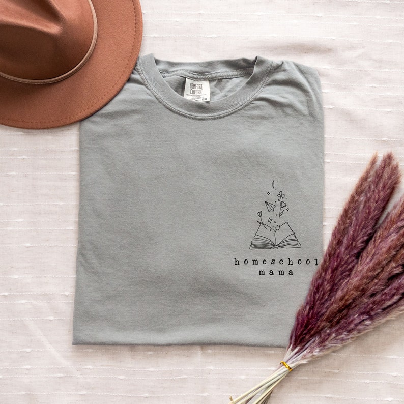 May include: A light gray t-shirt with a graphic of an open book with paper airplanes and flowers coming out of it. The text "homeschool mama" is below the graphic.