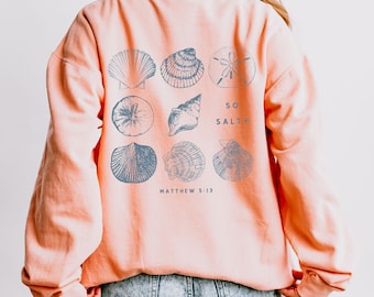 May include: A coral-colored sweatshirt featuring a collection of detailed seashell illustrations in shades of blue. The text "SO SALTY" and "MATTHEW 5:13" are printed on the back. The sweatshirt has a relaxed fit.