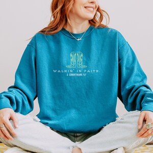 May include: A teal blue sweatshirt with a yellow and white graphic of two cowboy boots and the text "Walkin' in Faith. 2 Corinthians 5:7".
