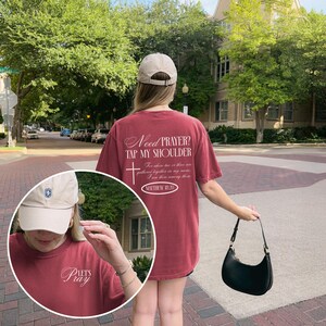 May include: A maroon t-shirt with the text "Need Prayer? Tap My Shoulder" and a cross graphic. The shirt also features the text "Matthew 18:20". A beige baseball cap and a black handbag are also visible.