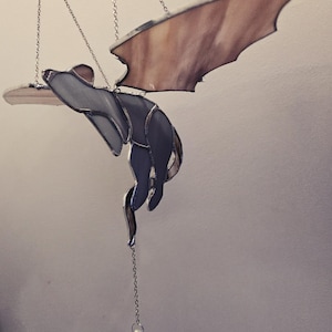 Può includere: Un drago in vetro colorato con le ali spiegate, appeso a una catena d'argento con un prisma di cristallo che pende sotto. Il drago è grigio e le ali sono di colore marrone chiaro.