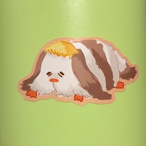 May include: A sticker featuring a cartoon creature with a white body, brown and white stripes, and a yellow tuft of hair. The creature has a sad expression with small eyes and a small orange nose. The sticker is on a light green background.