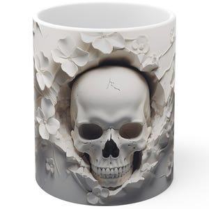 May include: White ceramic mug featuring a detailed, sculpted skull design. The skull is surrounded by white floral accents and set against a gray and white background. The mug has a standard cylindrical shape.