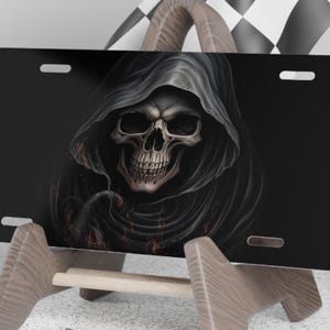 May include: Black license plate featuring a detailed illustration of a skull wearing a hooded cloak. The skull has a realistic appearance with visible teeth and eye sockets. The background is dark, enhancing the skull's features.