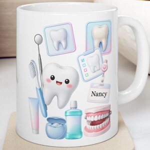 Puede incluir: Taza de cerámica blanca con ilustraciones de temática dental. Incluye un diente sonriente, cepillo de dientes, espejo dental, pasta de dientes, hilo dental, enjuague bucal y una dentadura. El nombre "Nancy" también se muestra.
