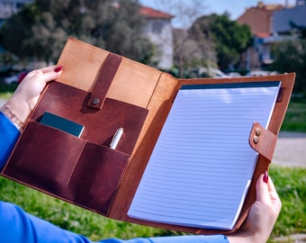 May include: A brown leather portfolio with a notepad and pen inside. The portfolio has a flap with a strap and a pocket on the inside.