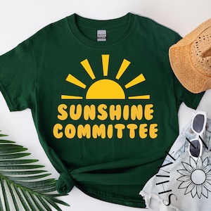 Op de afbeelding: Donkergroen t-shirt met een gele zon en de tekst "SUNSHINE COMMITTEE". De afbeelding bevat een strohoed, zonnebril en een spijkerbroek short.