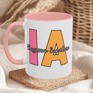 Op de afbeelding: Witte keramische mok met een roze binnenkant en handvat. De mok heeft de letters "IA" in roze en oranje, met de woorden "Classroom Superhero" in een cursief lettertype. De mok staat op een kurken onderzetter.
