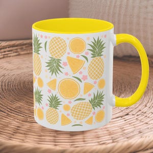 May include: A white ceramic mug with a yellow rim and handle. The mug features a repeating pattern of pineapples, lemons, and flowers in yellow, green, and pink.