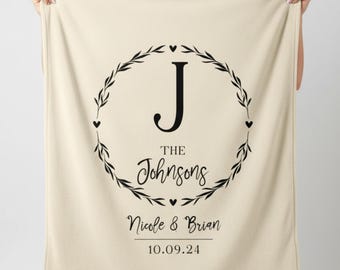 Puede incluir: Manta color crema con un diseño de corona circular negra. La corona rodea la letra "J" y las palabras "THE Johnsons". Debajo, los nombres "Nicole & Brian" y la fecha "10.09.24" están impresos en negro. La manta está sostenida por manos invisibles.
