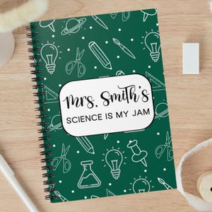May include: A spiral-bound notebook with a dark green cover featuring white science-themed illustrations. The notebook has a white rectangular label with the text "Mrs. Smith's SCIENCE IS MY JAM" in black script and block letters.