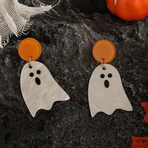 Puede incluir: Un par de pendientes de temática de Halloween con dijes de fantasmas blancos con ojos y bocas negras, que cuelgan de discos redondos cubiertos de purpurina naranja.