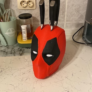 Puede incluir: Portacuchillos en forma de cabeza de Deadpool rojo y negro con cinco cuchillos plateados insertados en la parte superior.