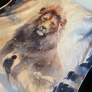 Op de afbeelding: Een aquarel schilderij van een leeuw op een beige achtergrond met blauwe en zwarte accenten. De leeuw kijkt naar voren met zijn mond dicht en zijn ogen open. Het schilderij heeft een zachte, dromerige uitstraling.