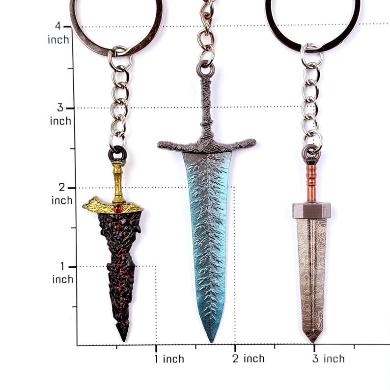 May include: Three metal sword keychains hanging from rings. The left keychain is gold and dark red, the center is blue and silver with a textured blade, and the right is copper and silver with a patterned blade. A ruler shows they are under 3 inches.