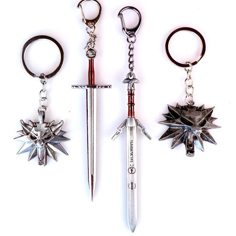 May include: Four metal keychains featuring two silver wolf head medallions and two sword pendants. The swords have red accents on the hilts and engraved markings on the blades. Each piece is attached to a silver-toned split ring by a short chain.