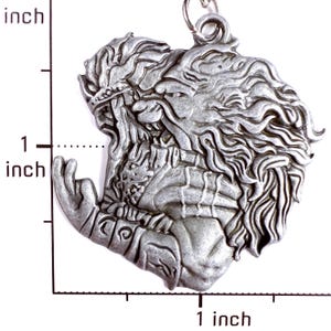 May include: A silver-toned metal pendant featuring a relief sculpture of a figure with long flowing hair and a beard, wearing a crown and armor. The pendant is positioned next to a ruler showing a height of approximately 1.5 inches.