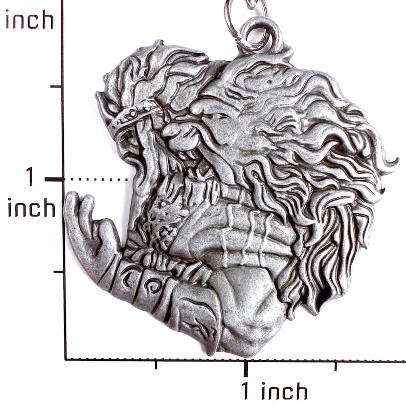 May include: A silver-toned metal pendant featuring a relief sculpture of a figure with long flowing hair and a beard, wearing a crown and armor. The pendant is positioned next to a ruler showing a height of approximately 1.5 inches.