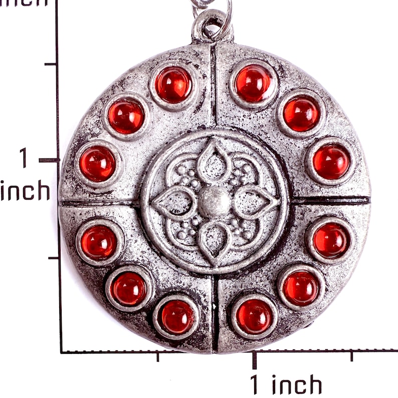 May include: A circular silver-toned pendant featuring a central embossed floral motif surrounded by ten small, round, translucent red cabochon stones. A ruler visible in the image indicates the pendant measures approximately 1.5 inches in diameter.