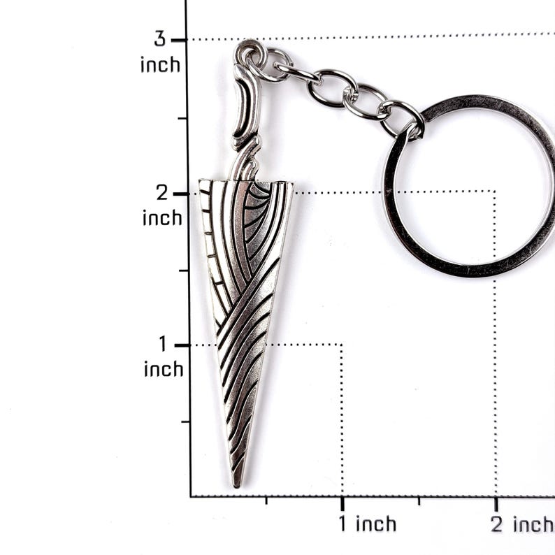 May include: A silver-toned metal keychain featuring a dagger-shaped pendant with an intricate engraved pattern of curved lines. The pendant measures approximately 2.5 inches in length, as indicated by the visible ruler markings in the image.