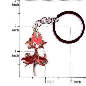 May include: A metal keychain featuring a character charm with a red and dark red color scheme, depicted with a pointed head and flowing lower section. The charm is attached to a silver-toned circular ring, with a visible scale showing a height of 1.5 inches.