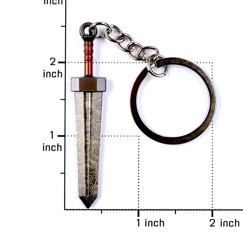 May include: A metal sword keychain featuring a dark blade with a gold-toned wavy pattern, a copper-colored handle, and a circular ring. The sword measures approximately 2 inches in length as shown against the visible imperial measurement scale.