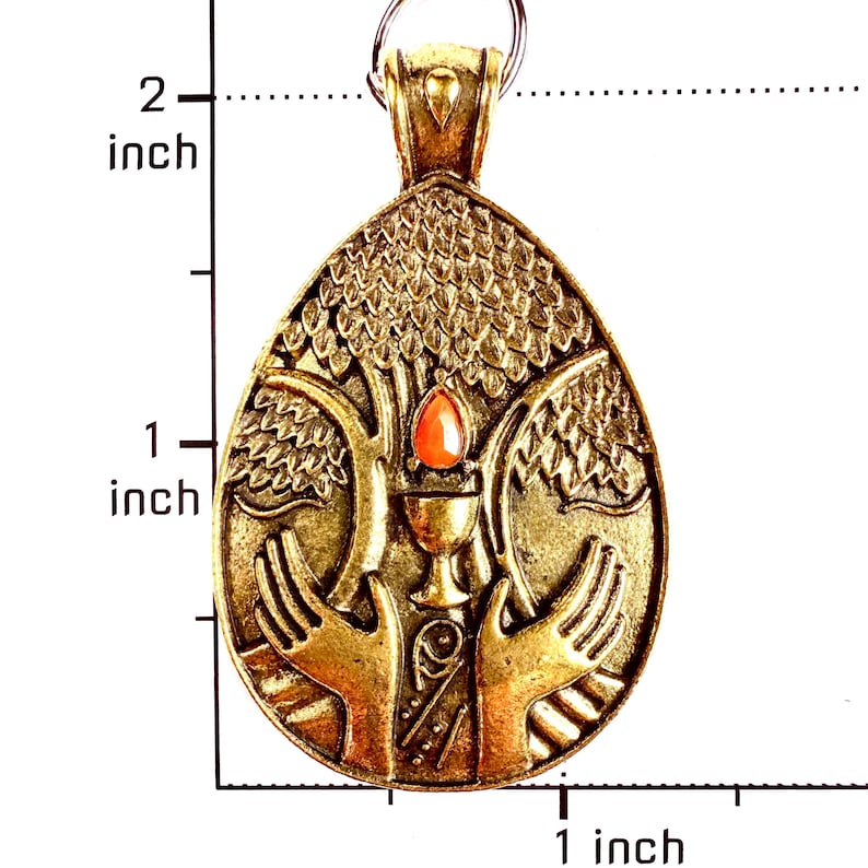 May include: A gold-toned metal pendant shaped like a teardrop, featuring a relief design of two hands cradling a chalice beneath a tree. A small orange stone is set above the chalice. A ruler indicates the pendant is approximately 2 inches tall.