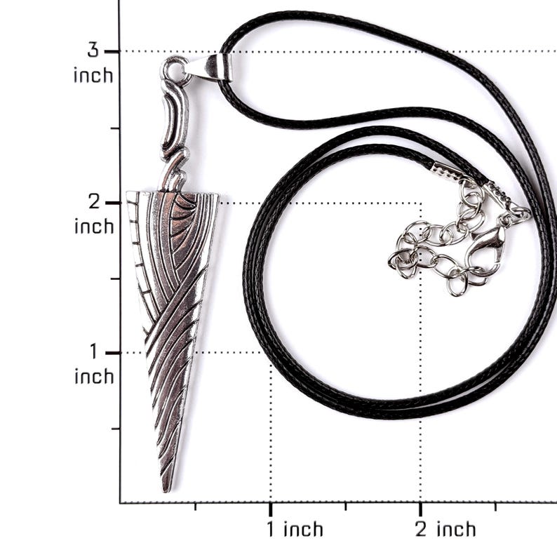 May include: A silver-toned metal pendant shaped like a stylized dagger with intricate engraved lines, suspended from a black cord necklace. A ruler overlay indicates the pendant length is approximately 2.5 inches and the width is 0.75 inches.