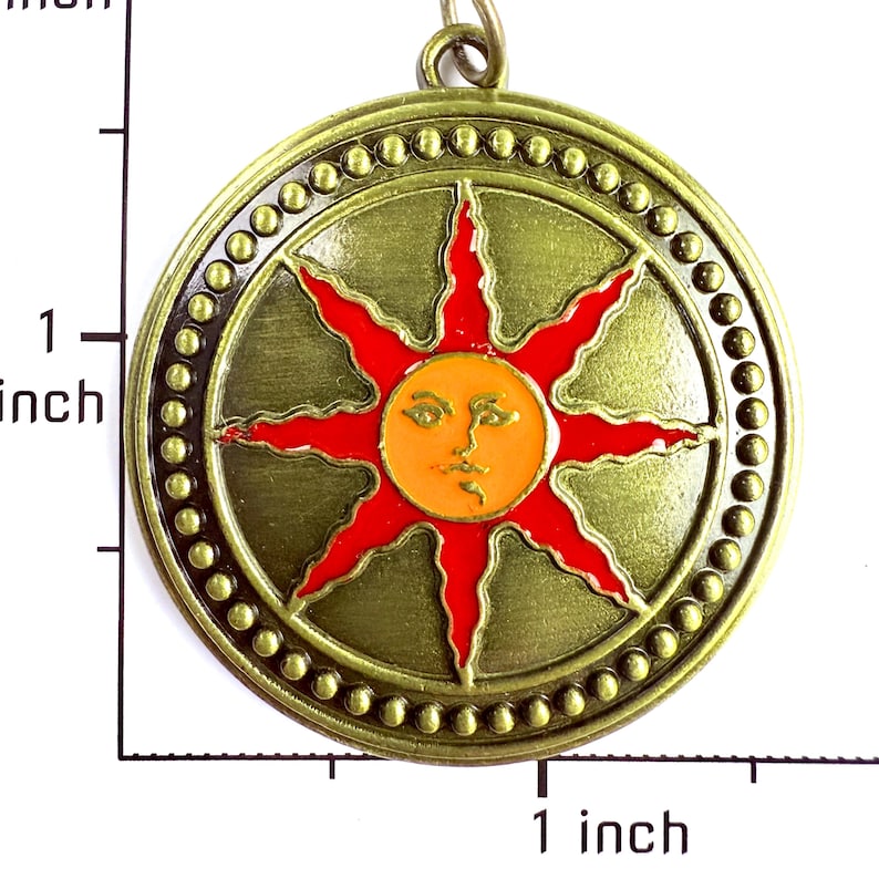 May include: A round brass-toned pendant featuring a central orange sun face with a red eight-pointed starburst design. The border has a beaded texture. A ruler visible on the left and bottom indicates the pendant is approximately 1.5 inches wide.