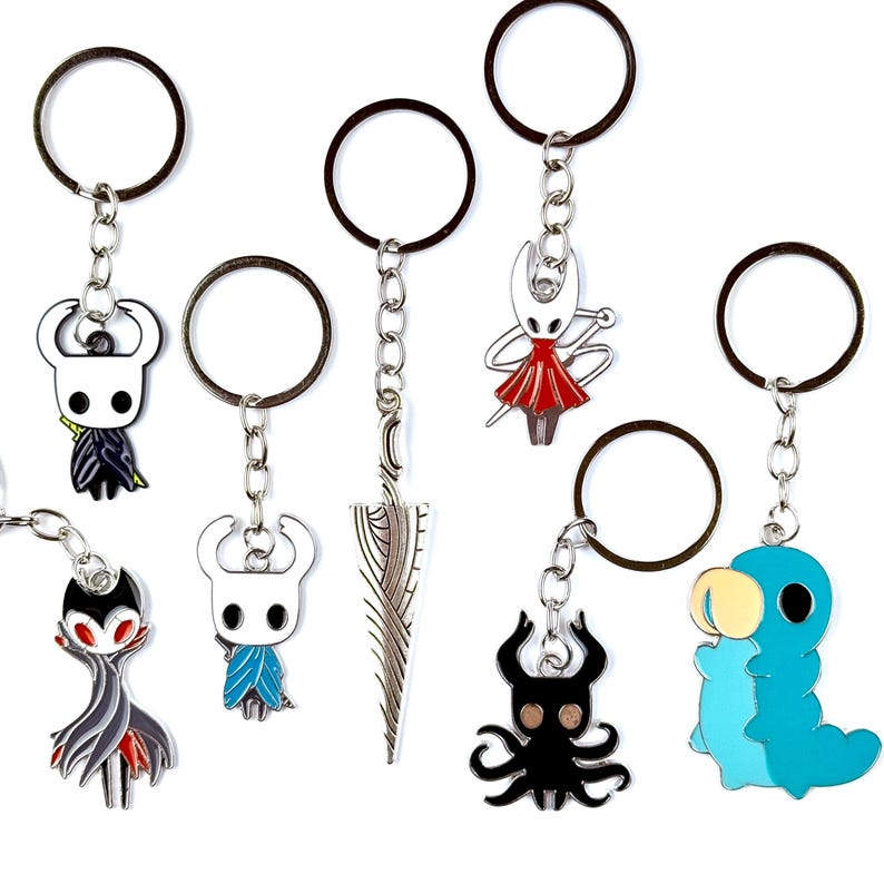 May include: A collection of seven metal keychains featuring various characters and a sword from the video game Hollow Knight. The charms include a white ghost figure, a red-cloaked character, a blue creature, a black multi-limbed figure, and a blue grub.