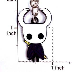 May include: A metal keychain featuring a white, horned character with black circular eyes and a dark purple cloak, positioned next to a ruler showing a height of approximately 1 inch. The character has a small yellow detail on its side.