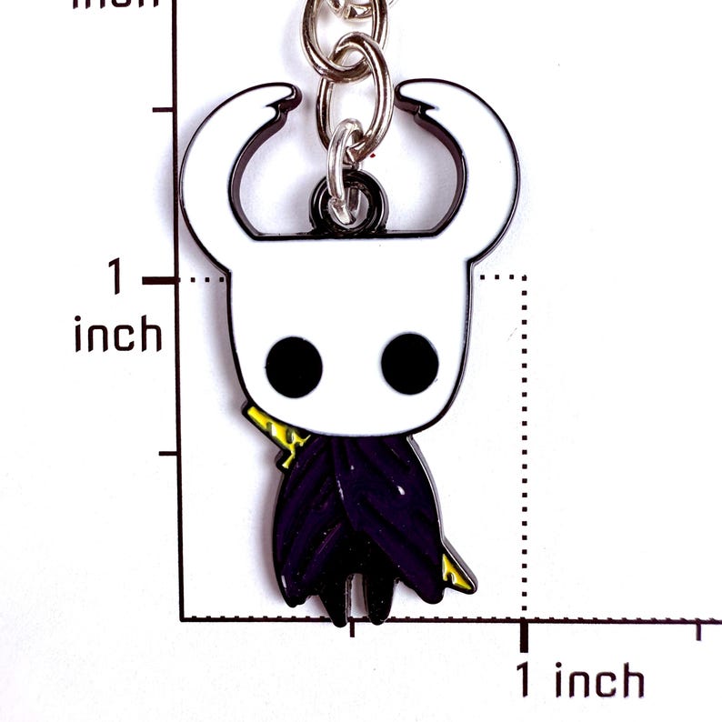 May include: A metal keychain featuring a white, horned character with black circular eyes and a dark purple cloak, positioned next to a ruler showing a height of approximately 1 inch. The character has a small yellow detail on its side.
